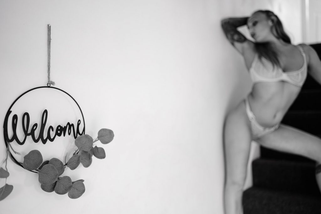 Black white photo of a woman in bra and pants stood on the stairs, she is blurred as the focus is on a Welcome sign in the foreground