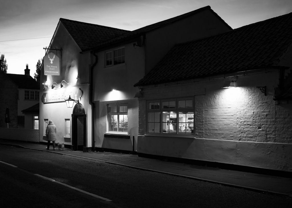 A black and white high contrast image of a village pub, its dark as night is falling. Pools of light are visible from the external lights of the pub and a woman is passing walking her dog.