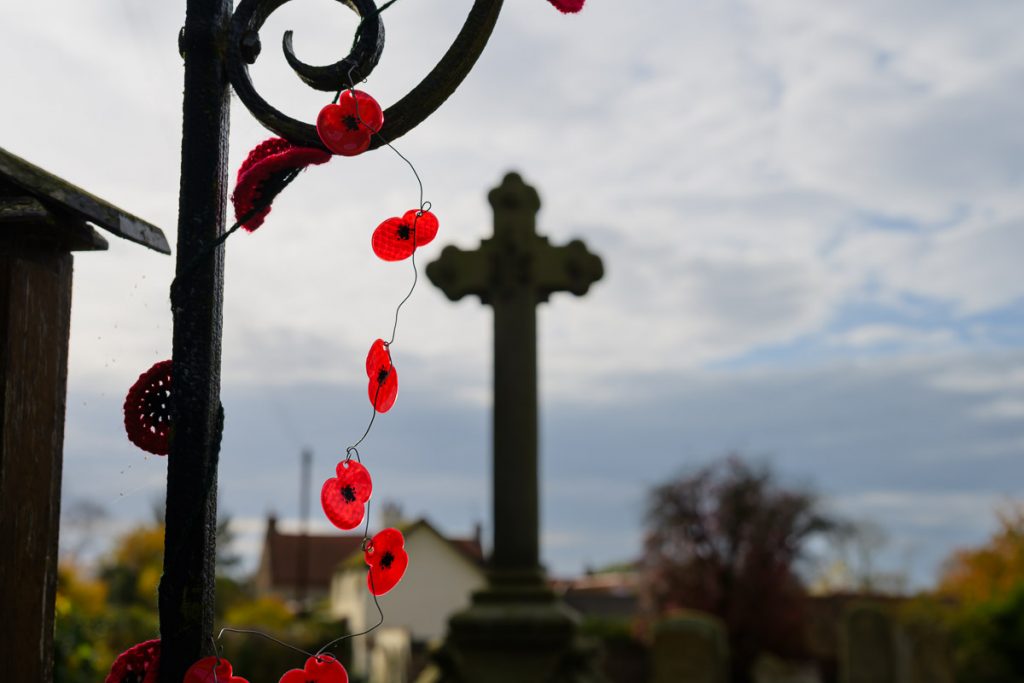 Some home made poppy flowers hanging from a gate post in a cemetery with a cross in the background. 