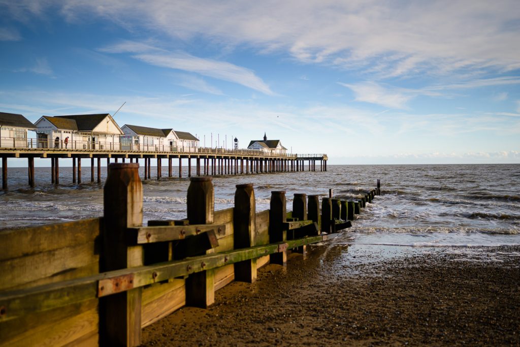 Picture of Southwold Pier disappearing into the sea. The white buildings on the pier can be seen and a wooden Groyne is seen in front mirroring the direction of the pier.