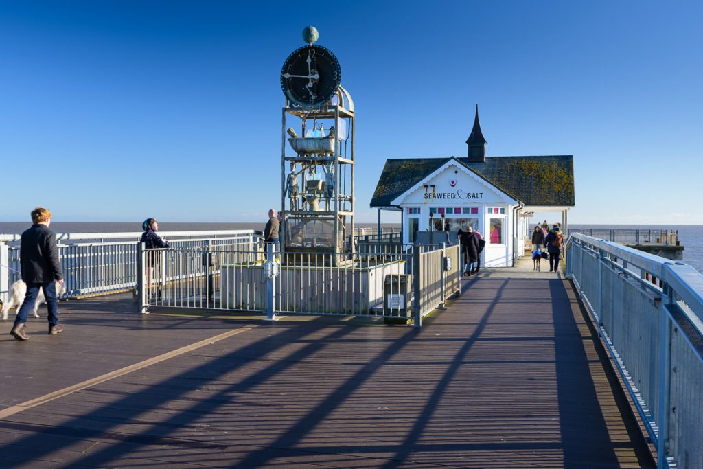 Picture of Southwold pier.  The cafe shop at the end can be seen, and people walking dogs on the pier.  A clock is central and the sky a clear blue.