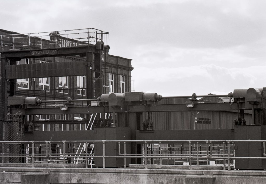 An old black and white film photography. The lifting gear of Stamp End Lock is in view with an old warehouse in the background.
