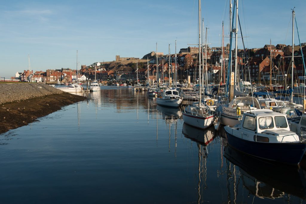 A view of the river leading to the harbour at Whitby.  The church next to the Abbey can be seen high up in the distance will lots of small boats moored on the right hand side.