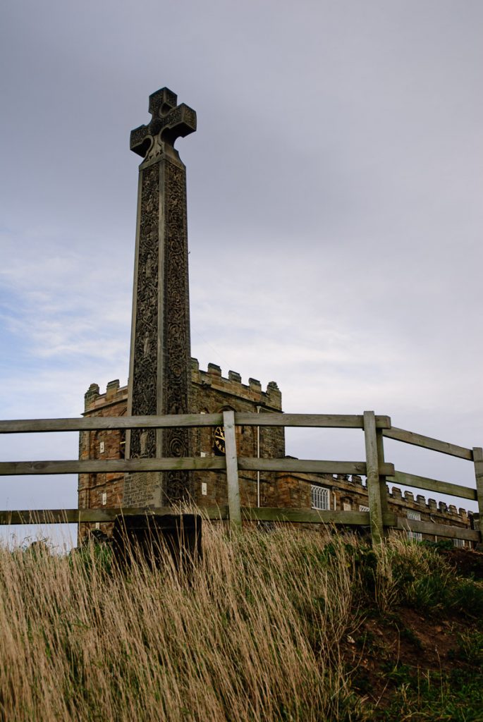 The old cross at Whitby in the foreground. There is a wooden fence in front of it and the church on the hill by the Abbey can be seen in the background.