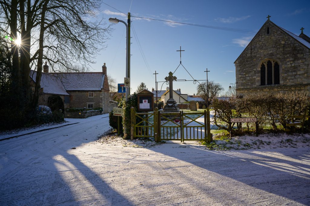 Winter scene.  Old church to the right, snow on the road and the sun can be seen coming though the trees on the left.