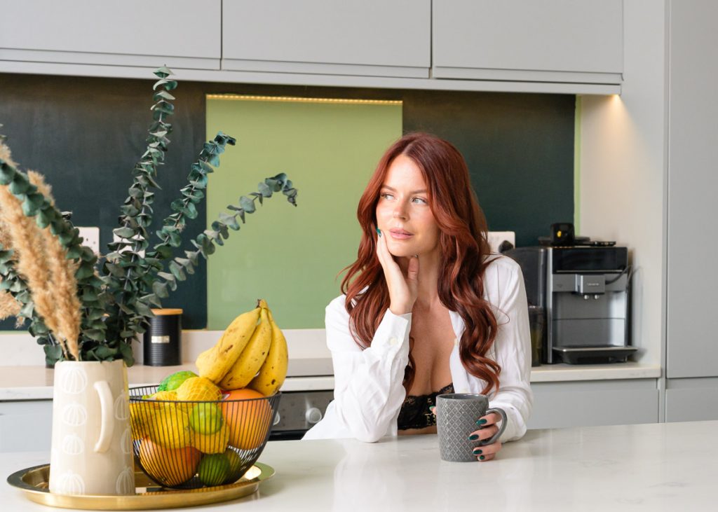 Red haired Amber sat in kitchen looking camera left. She is sat has one hand to her face and the other holding the coffee cup. She has an open white blouse with her black bra showing.