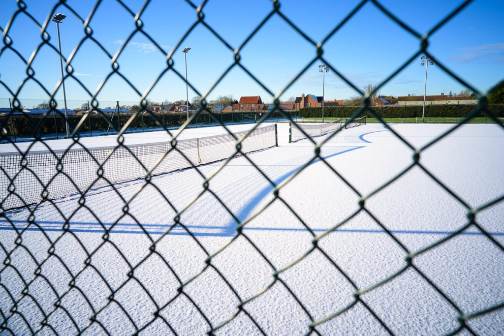 Photo through a wire fence of snow covered tennis courts. The sky is blue on a cold sunny day.