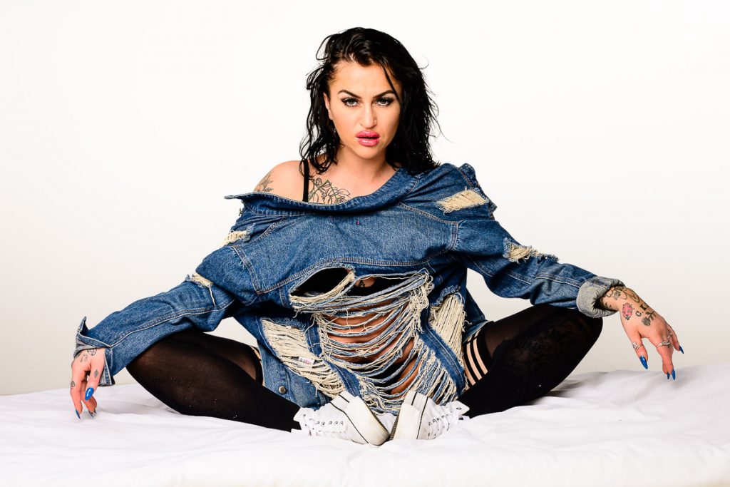 Model Rae with dark hair, sat on a shite sheet, wearing black bra and pants, black stockings, white trainers and a denim jacket reversed.