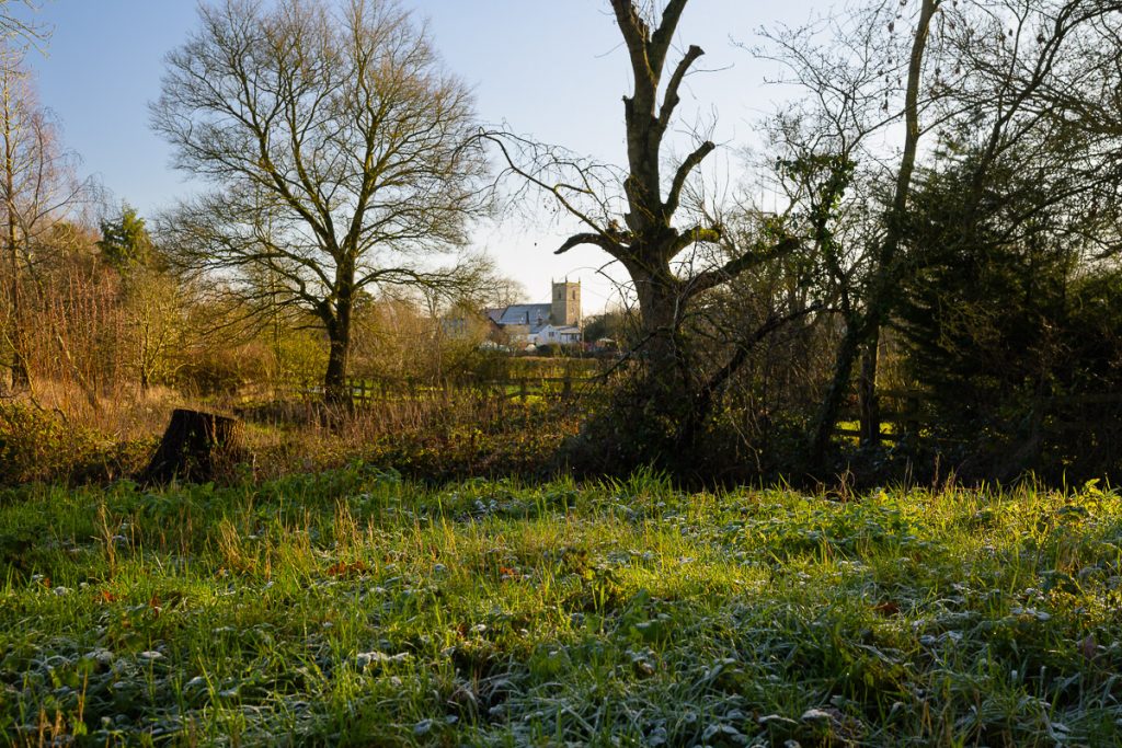 A photo through the bare winter trees of a small village Norman church.  In the grass, in the foreground there is still a small amount of snow, but the sky is blue and its a sunny day.