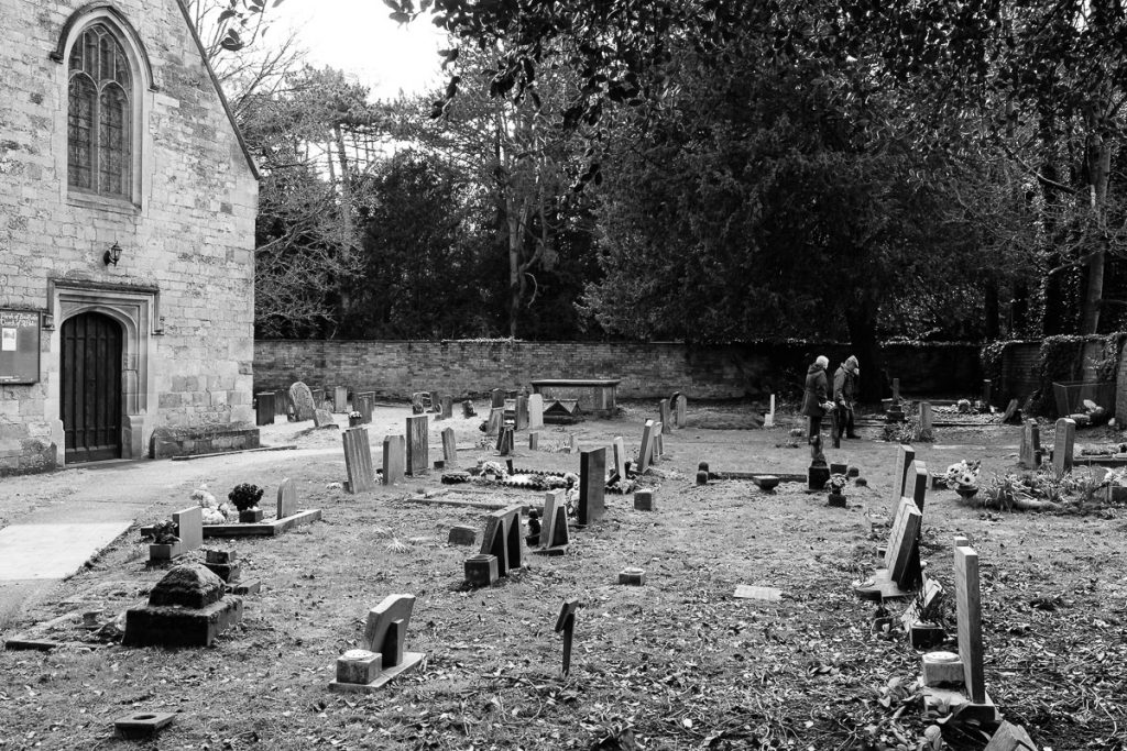 Black and White image of an old Church and grave yard. The entrance of St Helen's can be seen to the left with the extensive grave yard around it. Two figures in the distance look at one of the graves.