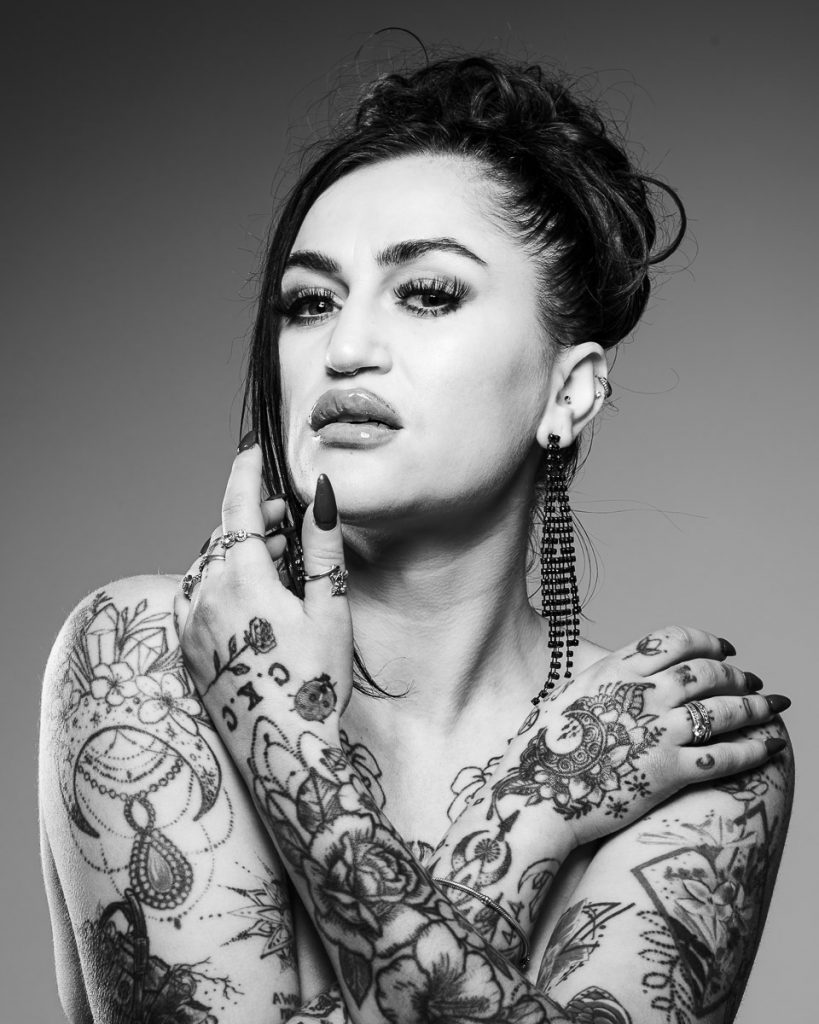 Black and white, implied topless portrait of a young female model who is heavily tattooed.  She had one hand to her face and is looking at camera.