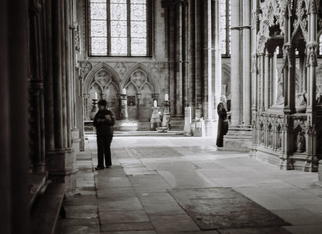 A slightly blurred photo inside Lincoln Cathedral.  A priest and a member of the public can be seen with logs of candles and a large window at the back.
