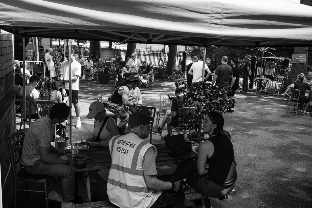 The out door food area, people sat at tables enjoying a drink and their food. The table at the front has some of the festival organisers on it with their laptops, keeping the online available beer list up to date.