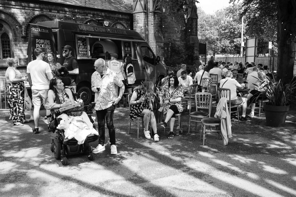 People enjoying the sun in the food area. A number of people are sat at tables enjoying the food and drink.