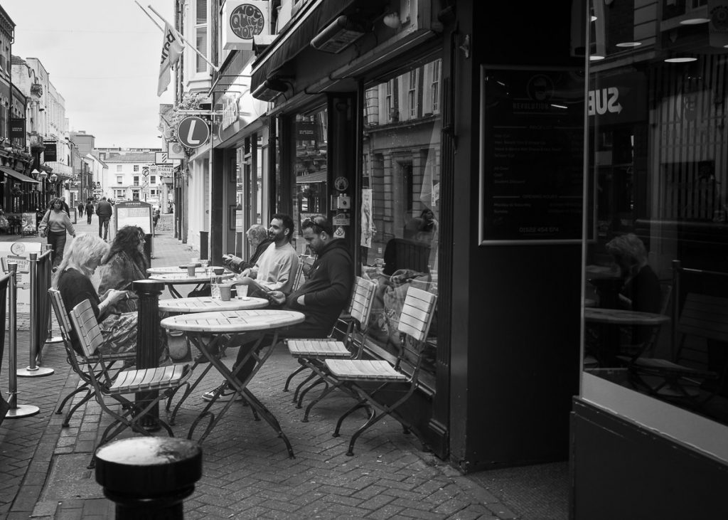 Tables outside a coffee shop, several of the tables of occupants drinking coffee and having a chat.