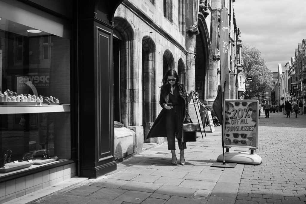 Woman walking past a Jeweller's shop.  She is concentrating on her phone, which she is looking down at in her hand.