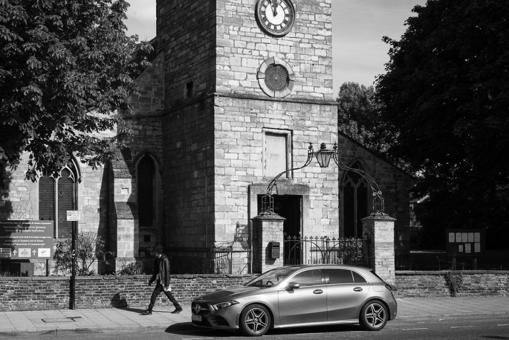 St Basil and St Paisios Greek Orthodox church in Lincoln. The clock down can be seen in the centre top of the tower and a man in a leather jacket walks by. A car is parked in front of the church.