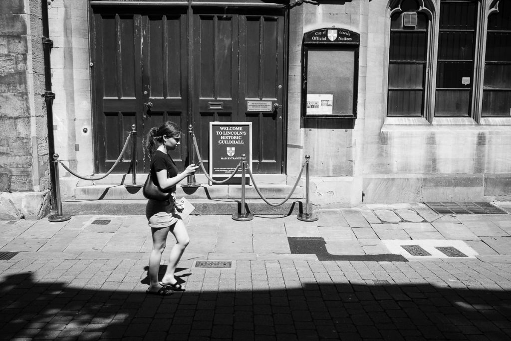 Entrance to Lincoln Guild Hall. Doors in the background, a young woman looking at her phone is walking past.