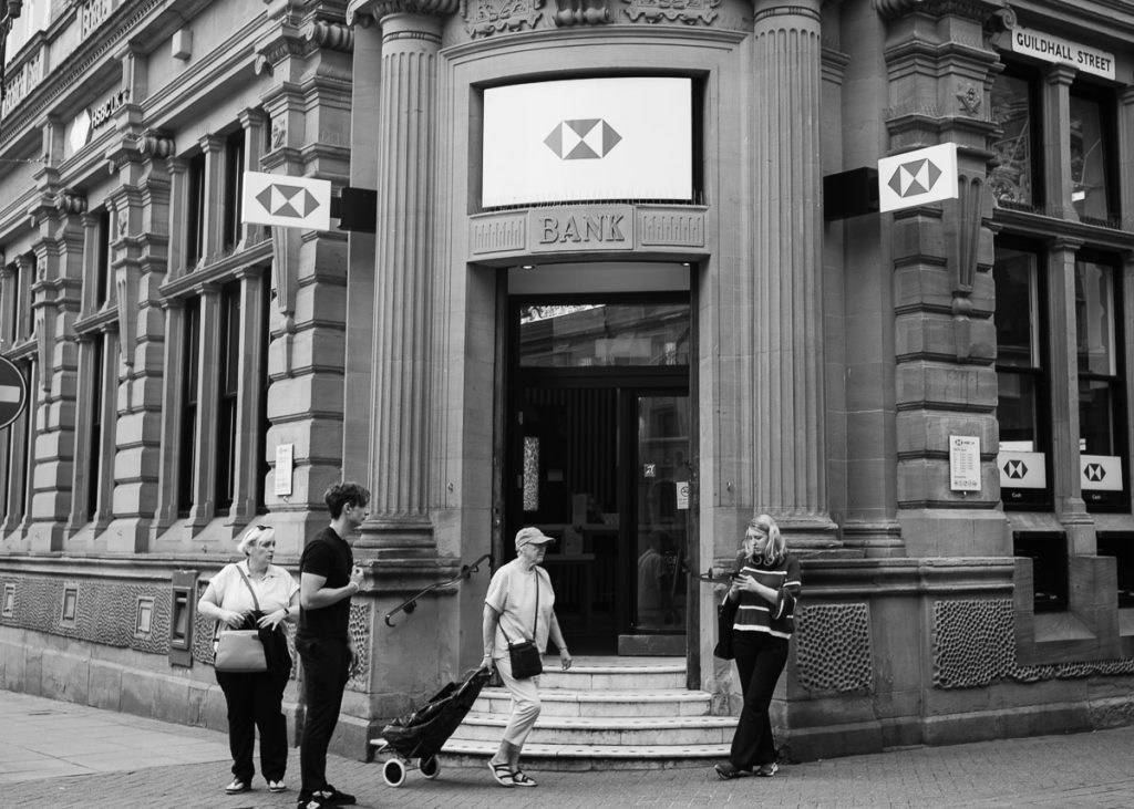 An old building that is still a high street bank in Lincoln.  In front are a few people passing, while one woman is checking her phone.