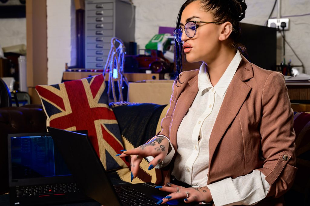 Black haired model with glasses, white blouse and pink jacket.  She is sat typing onto a laptop
