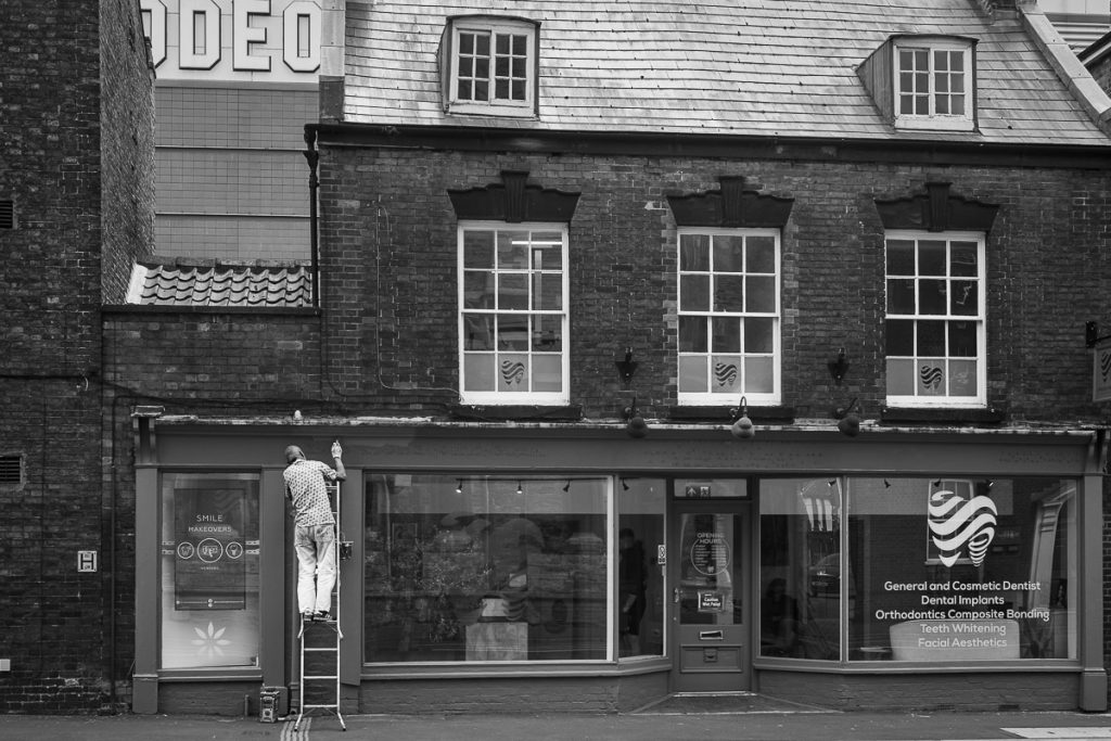Old shop front which is now a dentist, a modern building can just be seen.  A man in on a ladder doing some cleaning.