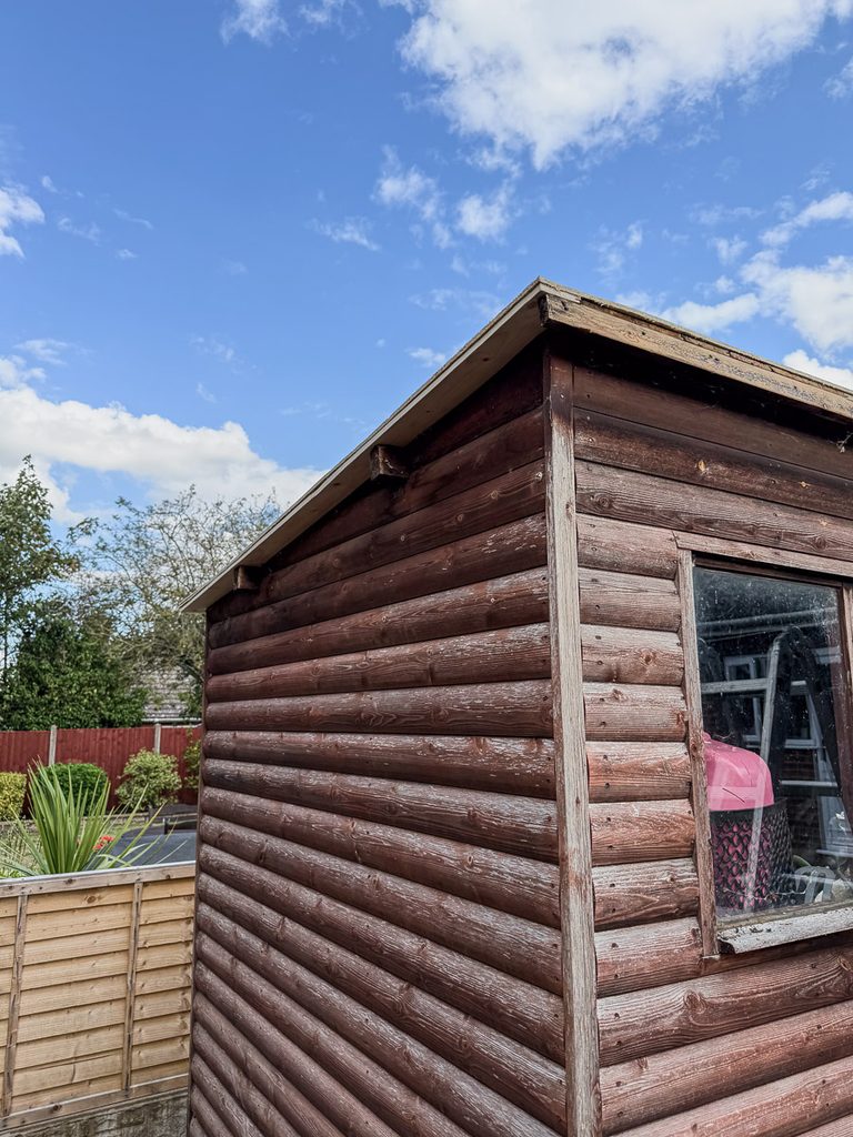Picture of a wooden she, the roof can be seen and a window,  The sky is bright blue with white clouds