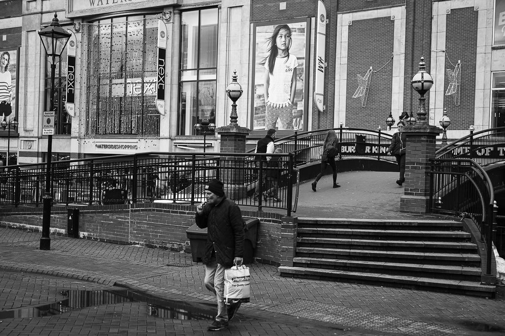 Picture of waterside Lincoln, large billboards can be seen in the background, a few people can be seen crossing the small bridge.  A man smoking in the foreground can be seen.