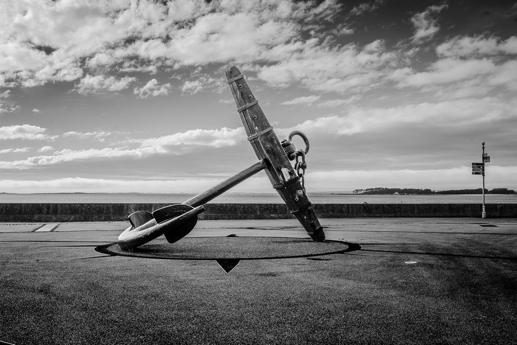 Old ships anchor on the sea front.  The image is black and white and there is a dramatic sky behind. 