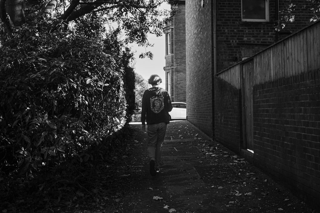 Woman walking away, large hedge to the left and a wall on the right. High contrast black and white